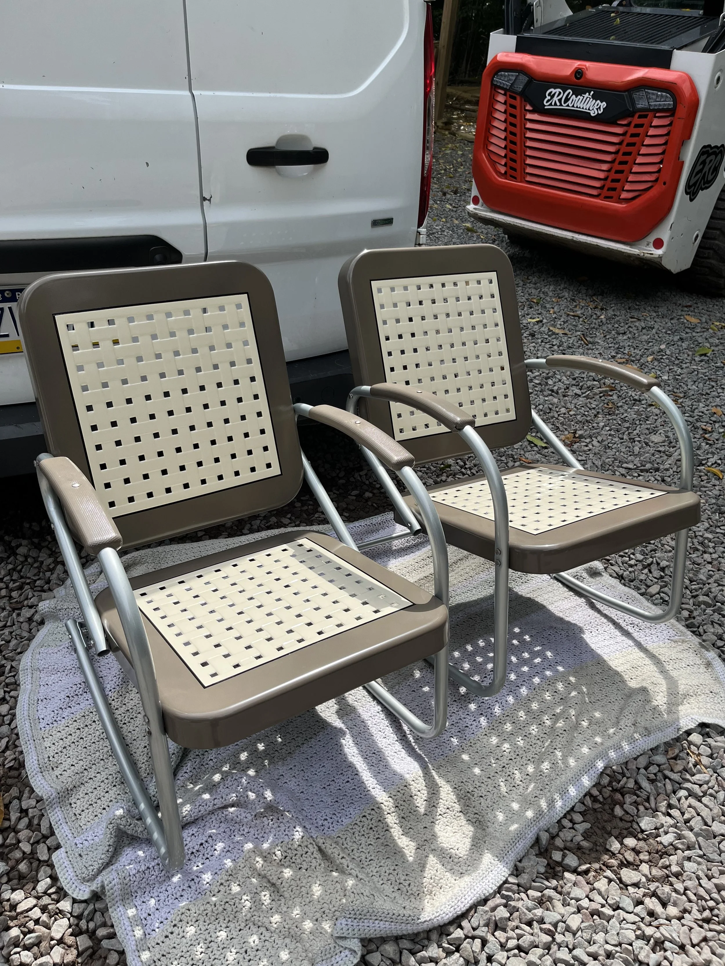 Two beige outdoor chairs with metal frames and woven plastic seats and backs, placed on a white carpet on gravel, with a white van and a red and white construction machine in the background.