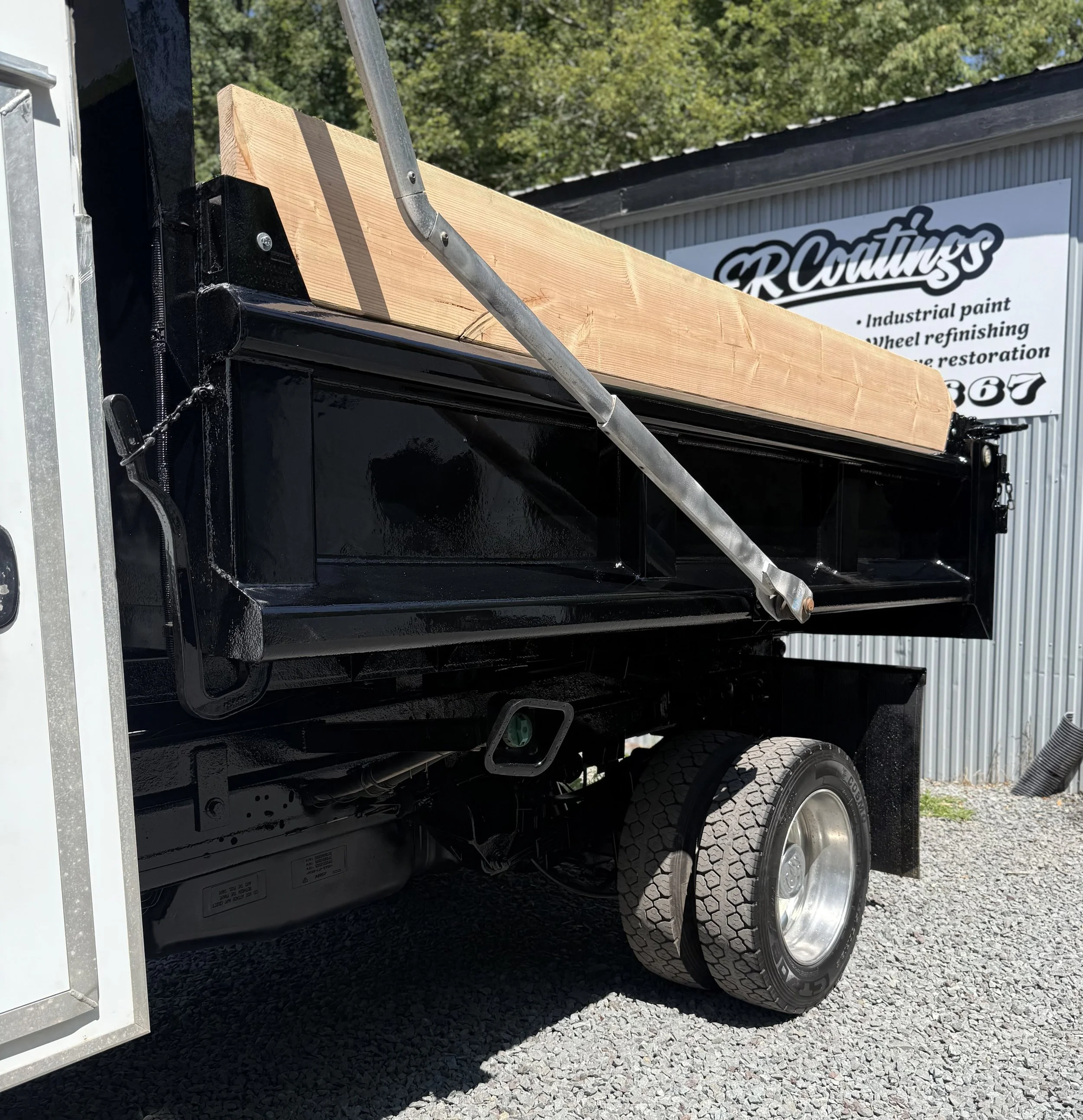 A black flatbed truck with a large wooden beam loaded on it, parked outside a building with a sign that reads 'R Coatings'.