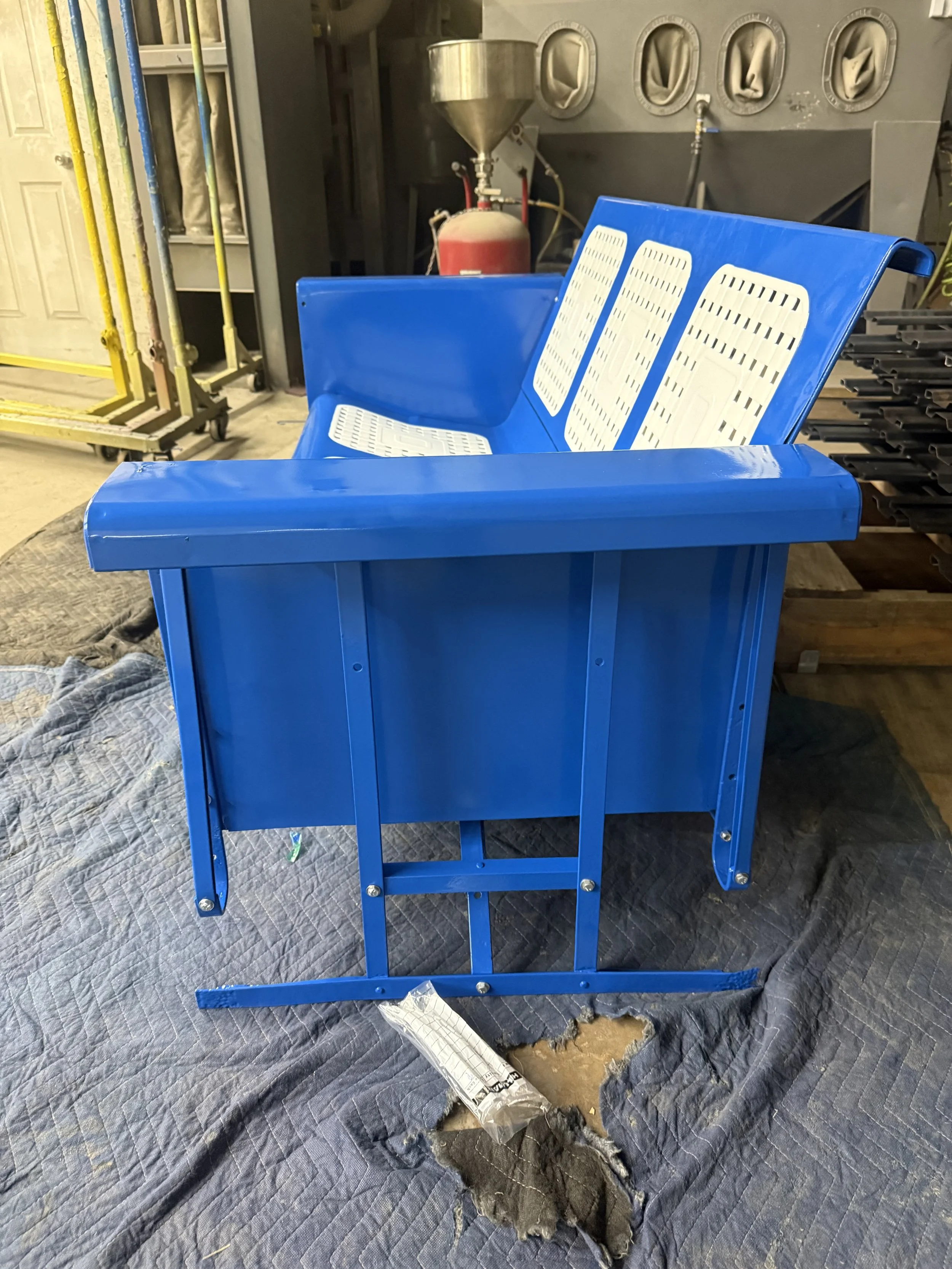 A bright blue stadium seat with white perforated metal slats on the backrest and seat, situated in an industrial or warehouse setting on a blue quilted blanket on a concrete floor.