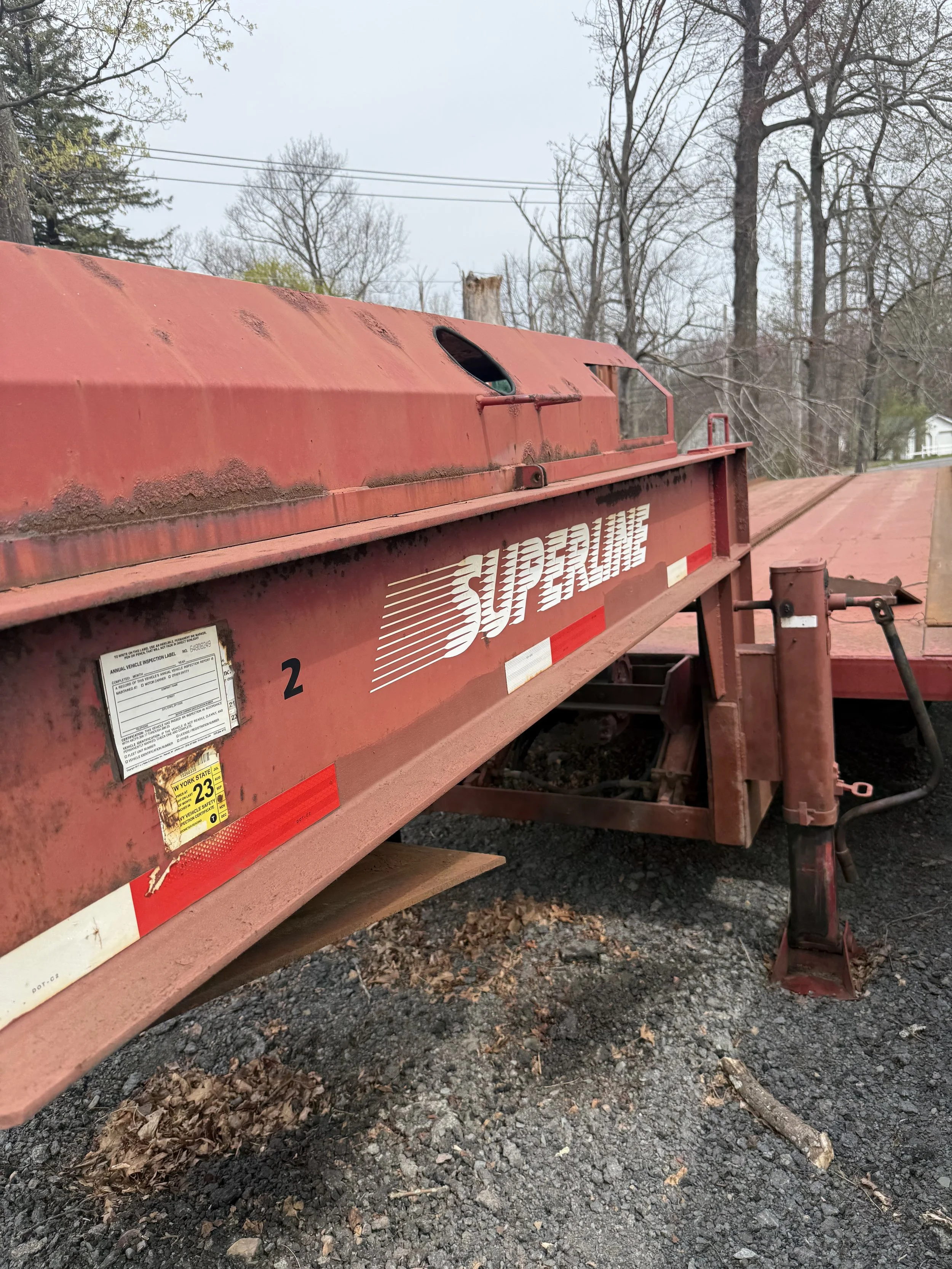 A red SUPRA TRAILER with white lettering and reflective red and white tape parked on gravel ground, with leaf litter and a few small branches nearby, and trees and power lines in the background.