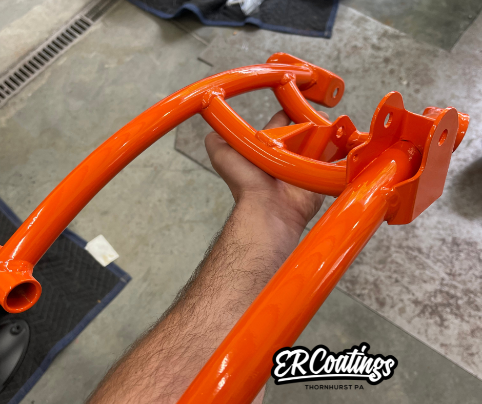 A person holding a freshly painted bright orange metal part, possibly a frame or a handle, in a workshop environment.