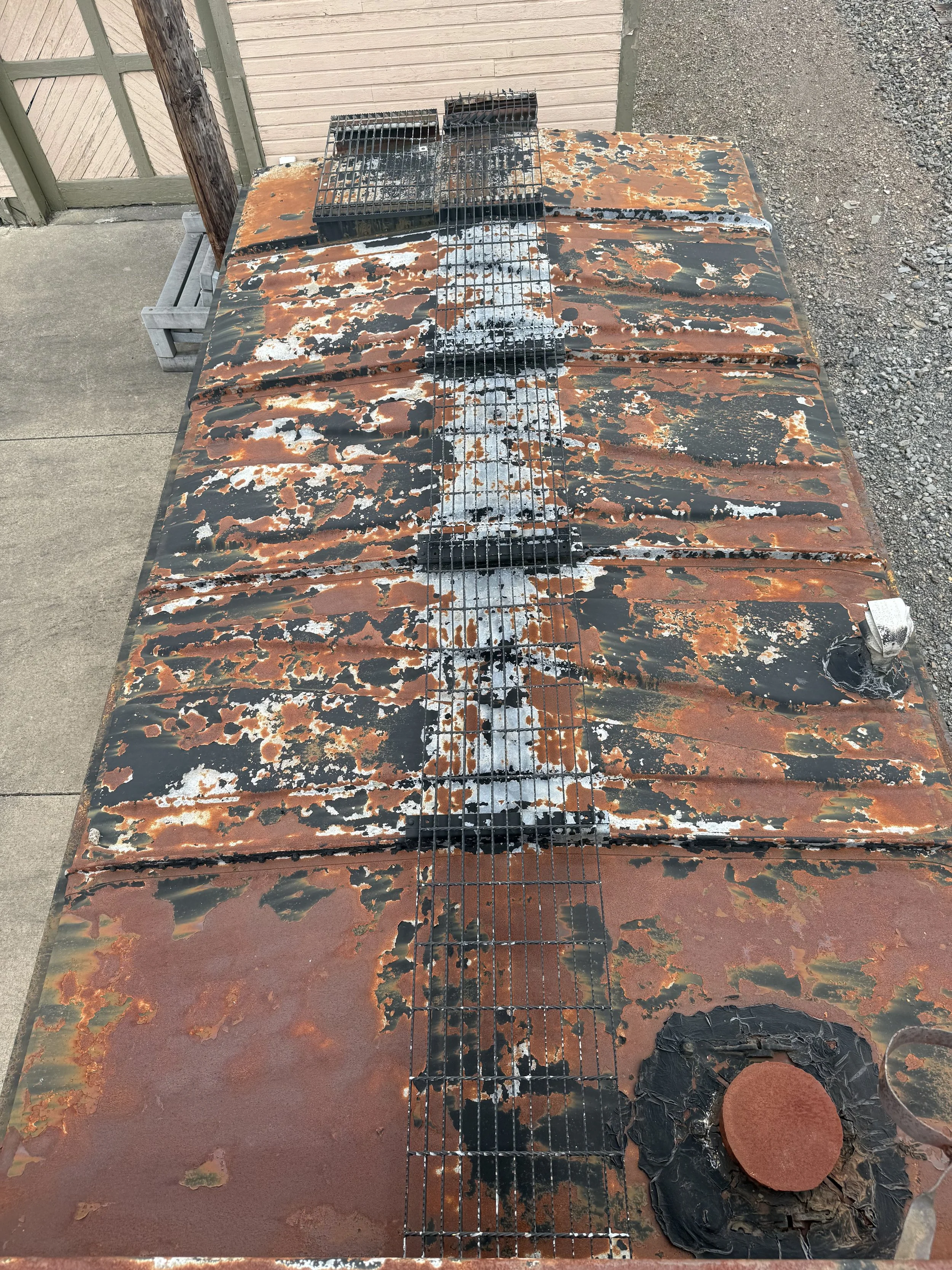 A rusted, weathered metal surface with peeling paint and a wire running down the center, with some blackened areas and objects on top.