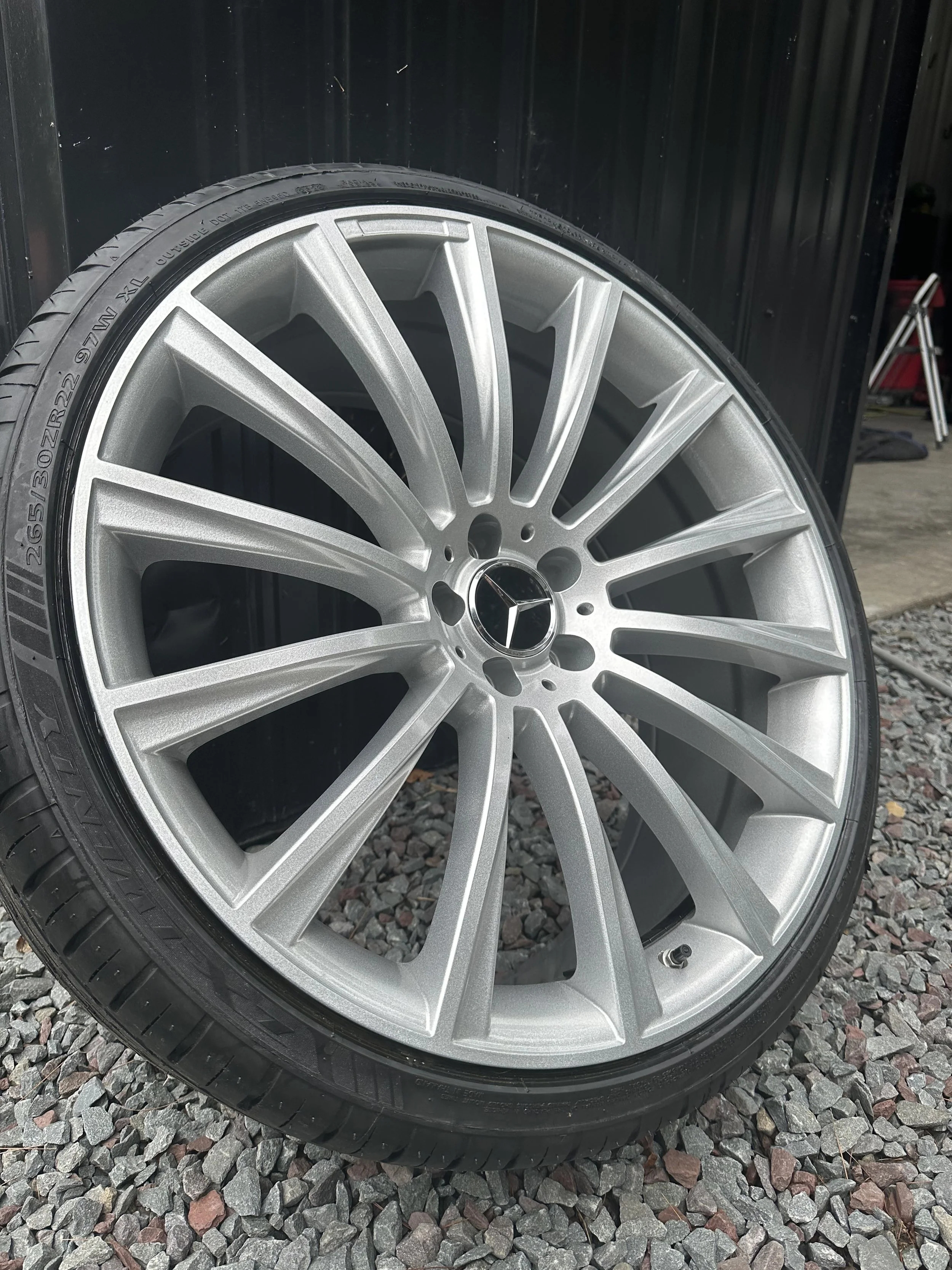 A silver Mercedes-Benz alloy wheel with a tire mounted on it, leaning against a black wall on a gravel surface.