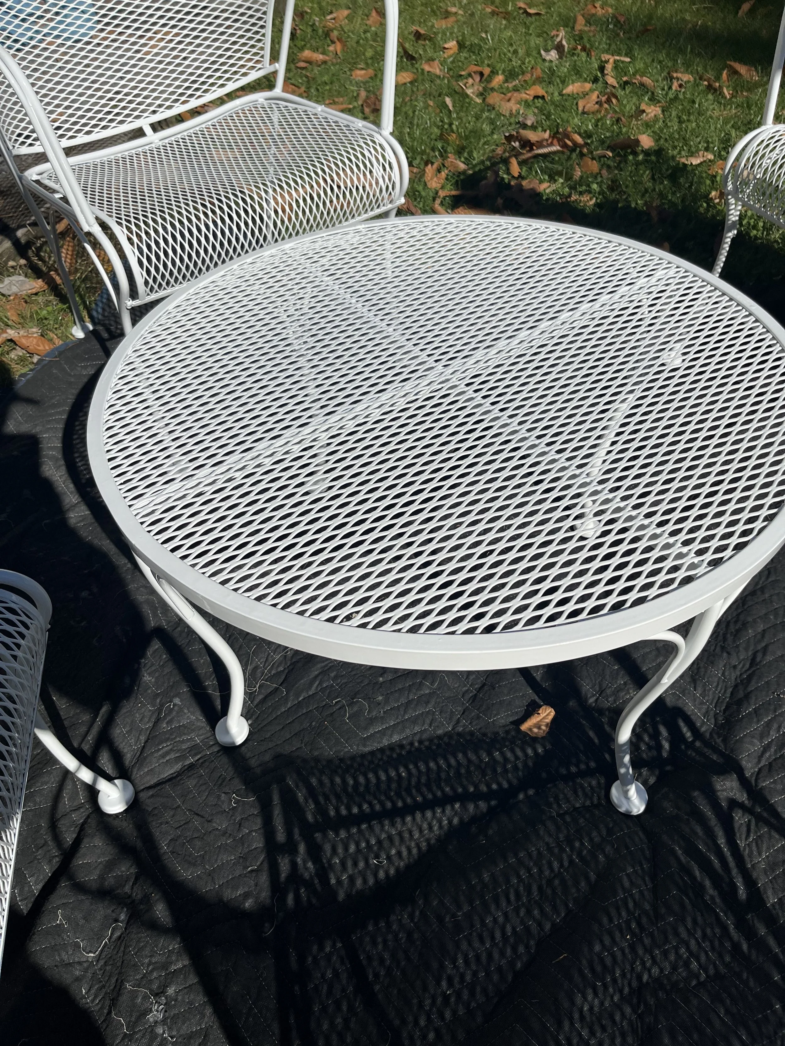White outdoor metal table and chairs on a black quilted fabric, with grass and fallen leaves in the background.