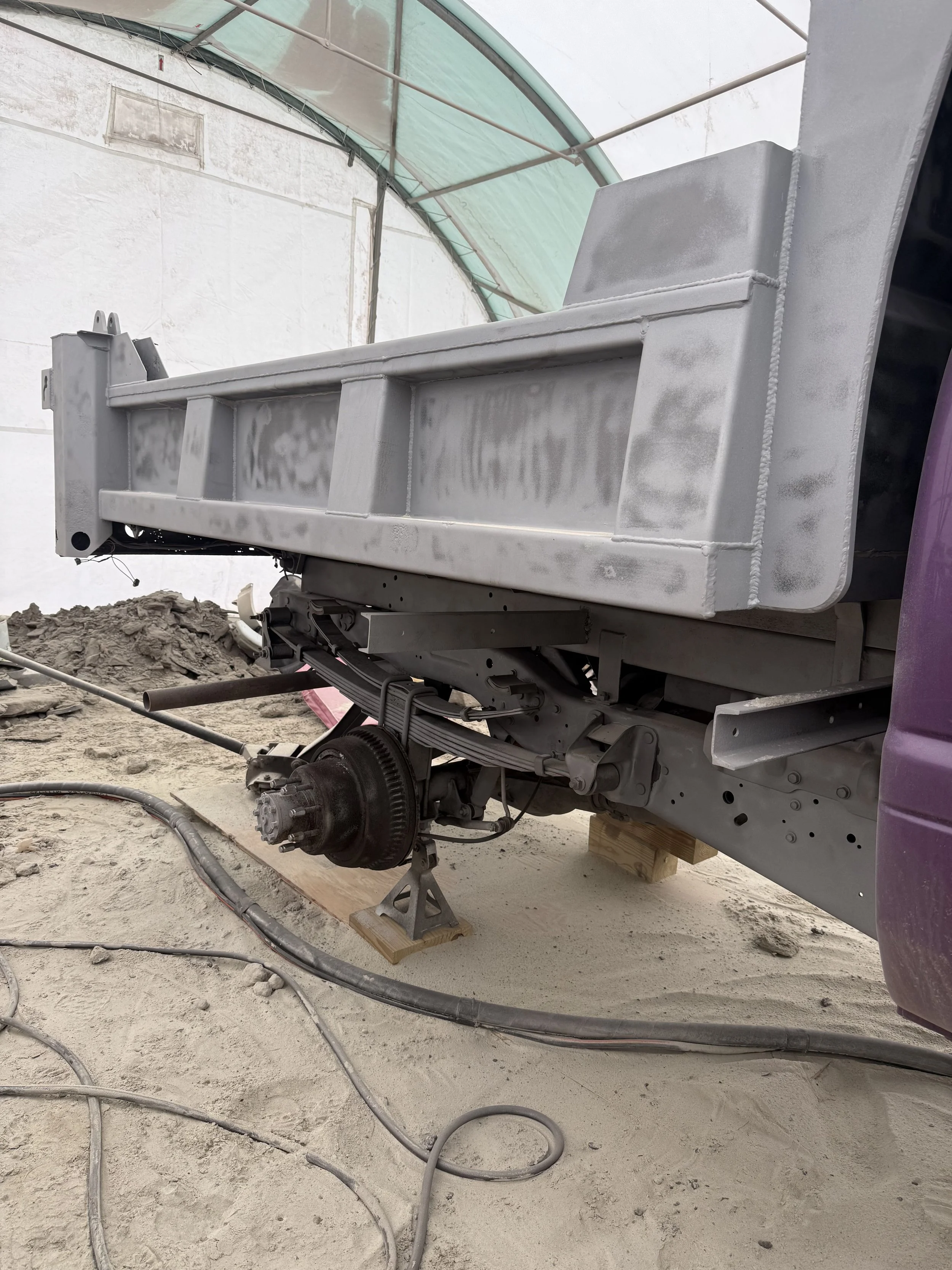 A truck chassis undergoing construction inside a covered workshop, with the wheel assembly removed, exposing suspension and axle components, supported on wooden blocks and concrete blocks, surrounded by construction tools and cables.