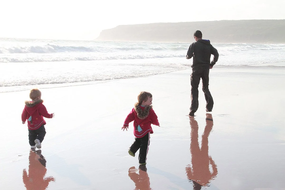 A person and two children walking on a beach with shallow water, waves, and cliffs in the background, during daylight.