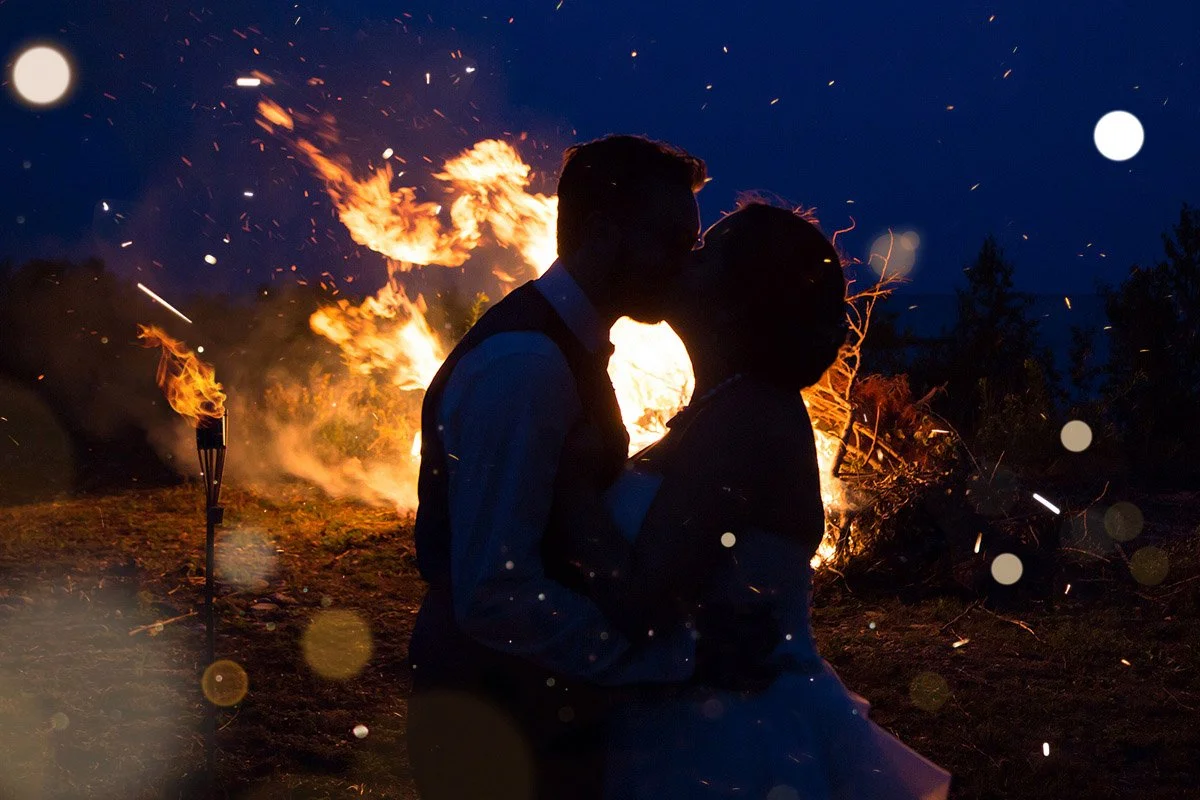 A silhouette of a couple kissing in front of a large outdoor fire at night.