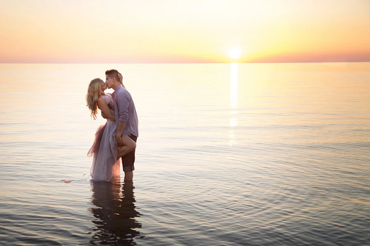 A couple kissing in the shallow water at sunset, standing in a calm lake with the sun setting in the horizon.