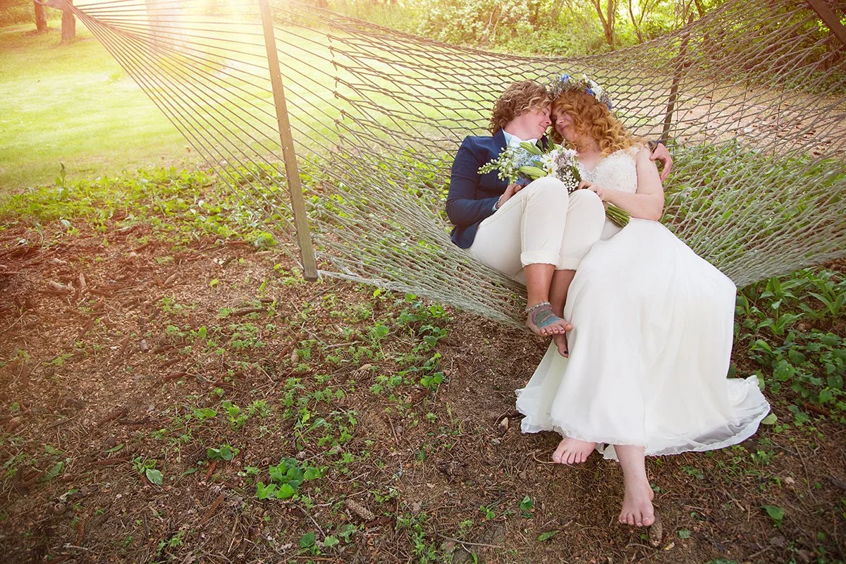 A couple dressed in wedding attire lying together in a hammock outdoors, surrounded by greenery, sharing an intimate and joyful moment.