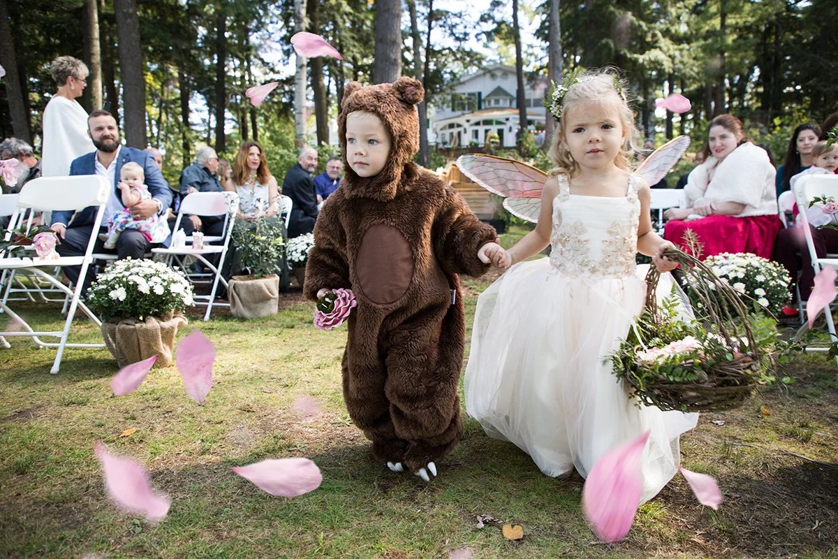 Two young children dressed as a bear and a fairy holding hands, walking down the aisle at an outdoor wedding ceremony surrounded by guests and flower decorations.