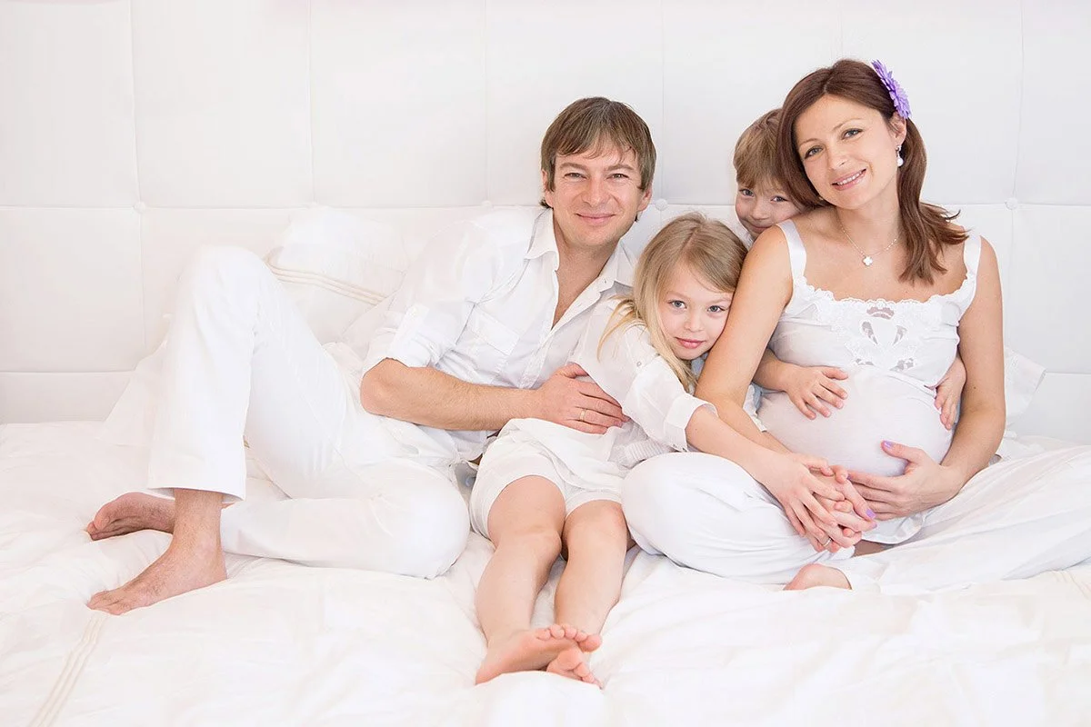 A pregnant woman and a family of four sitting on a bed, smiling, with three children hugging their mother.
