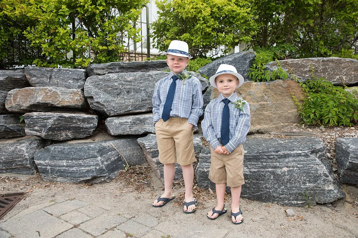 Two young boys dressed in matching outfits with plaid shirts, navy ties, khaki shorts, and white hats with blue bands, standing outdoors in front of a stone wall with greenery.