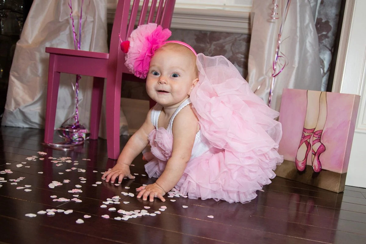 A baby girl dressed in a pink tutu and a big pink flower headband, crawling on a dark wooden floor amidst pink and white confetti. There is a pink chair, pink-themed decor, and a painting of ballet shoes in the background.