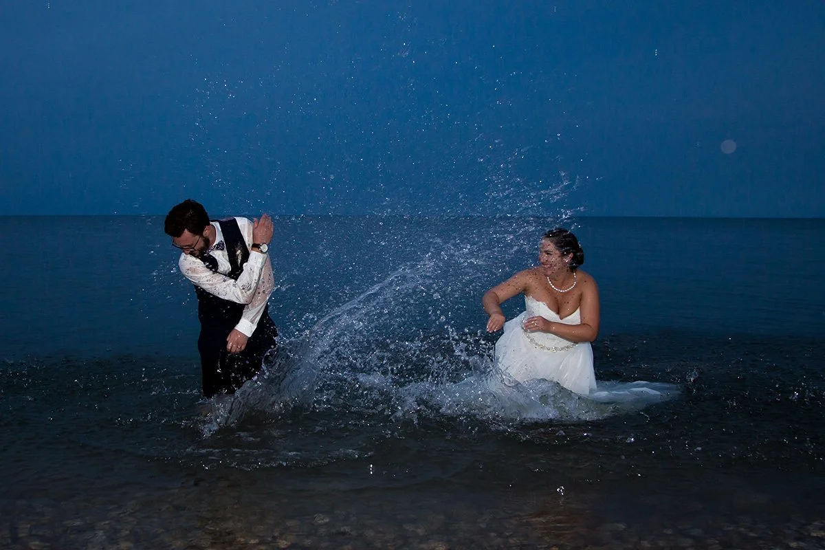 A man and woman in wedding attire playing in the ocean at dusk, with the woman smiling and the man ducking to avoid the water splash.