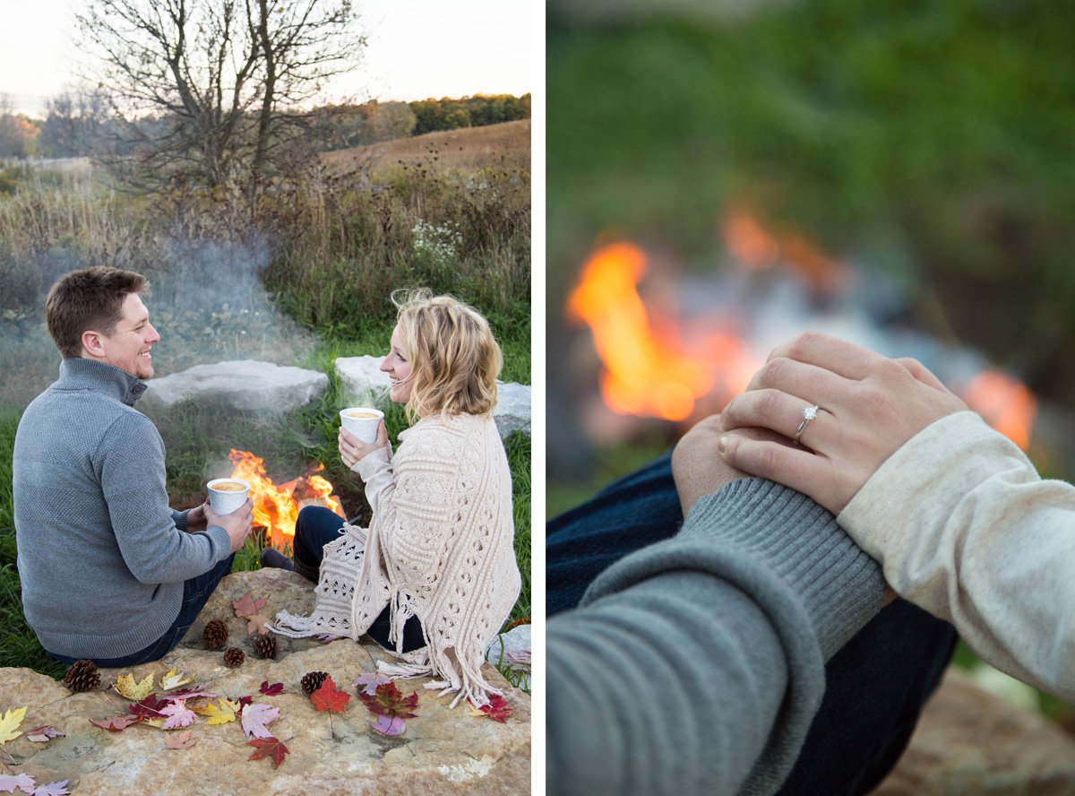 A couple sitting by a campfire outdoors, smiling and holding mugs, with autumn leaves scattered around. The woman is showing her engagement ring in a close-up shot as they hold hands with a fire in the background.