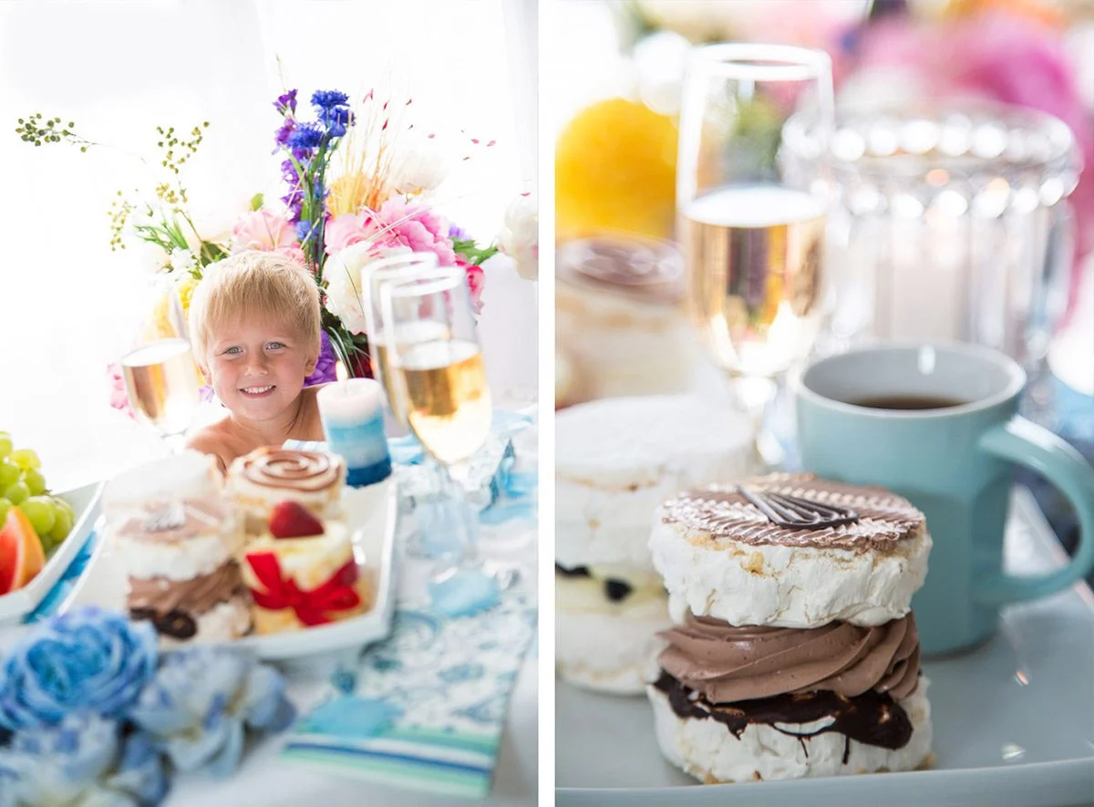 A children's birthday party with a smiling young boy. The table is decorated with colorful flowers, fruit, cupcakes, and desserts, with champagne glasses in the background.