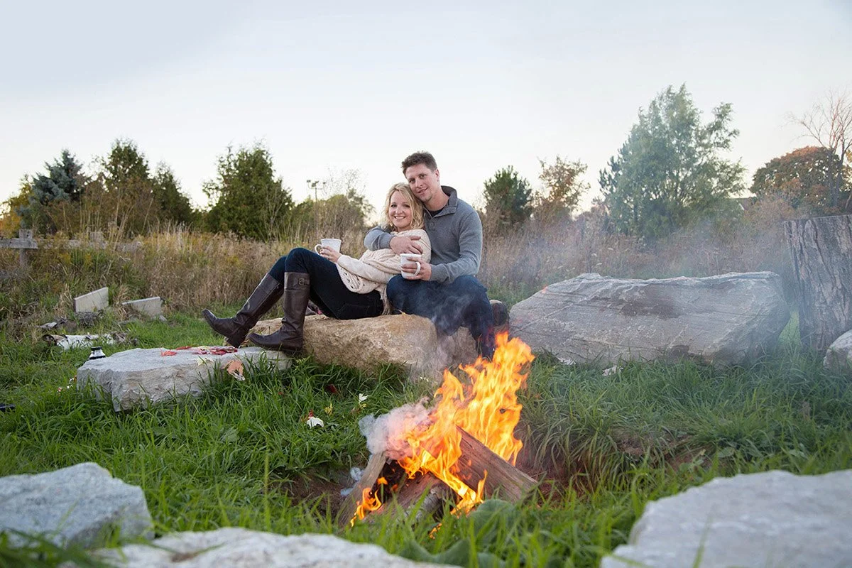 A smiling couple sitting on a large rock outdoors near a campfire, holding cups, with trees and a grassy field in the background.