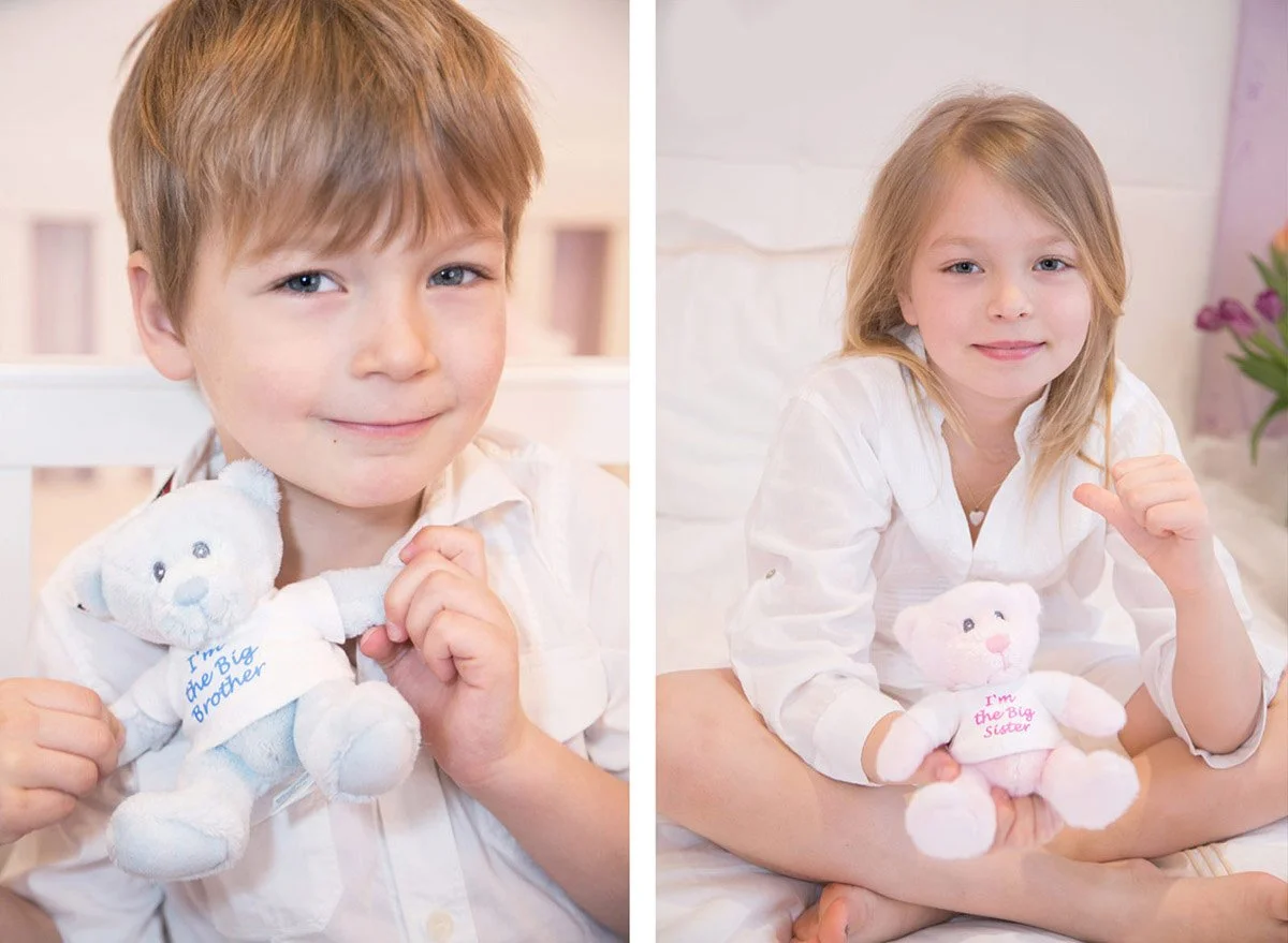 A young boy and girl each holding a plush teddy bear with embroidered text. The boy's bear says 'I'm the Big Brother' and the girl's bear says 'I'm the Big Sister'. They are sitting indoors, smiling at the camera.