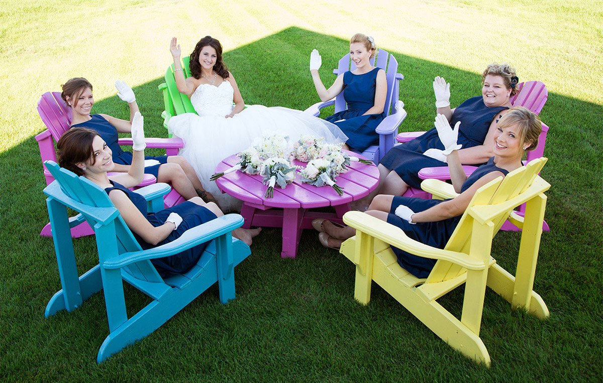 Bride in a white wedding dress and six bridesmaids in navy dresses sitting on colorful Adirondack chairs outside, waving at the camera, with a bouquet on the pink table in the center.