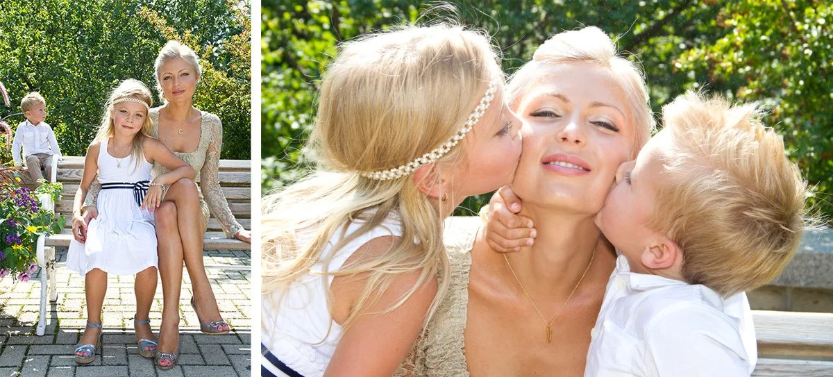 A woman sitting on a park bench with two young children, one girl and one boy, outdoors on a sunny day with trees in the background. The girl is sitting on the woman's lap, and the boy is kissing her on the cheek.