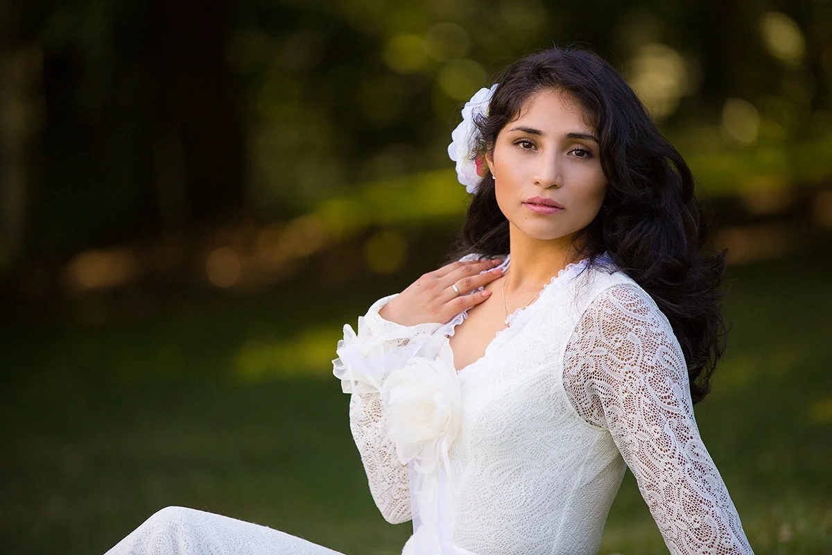 A young woman with curly dark hair wearing a white lace dress with floral accents, standing outdoors in a natural setting with trees and greenery in the background.
