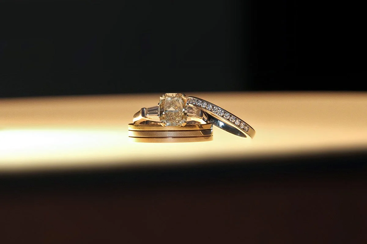 Close-up of three rings, one with a large central diamond, another with smaller diamonds, and the third plain, stacked on a reflective surface with a dark background.
