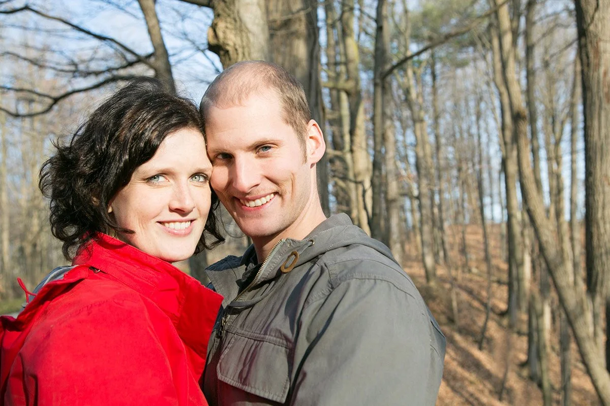 A smiling couple standing close together in a wooded park during daytime, surrounded by tall trees with bare branches.