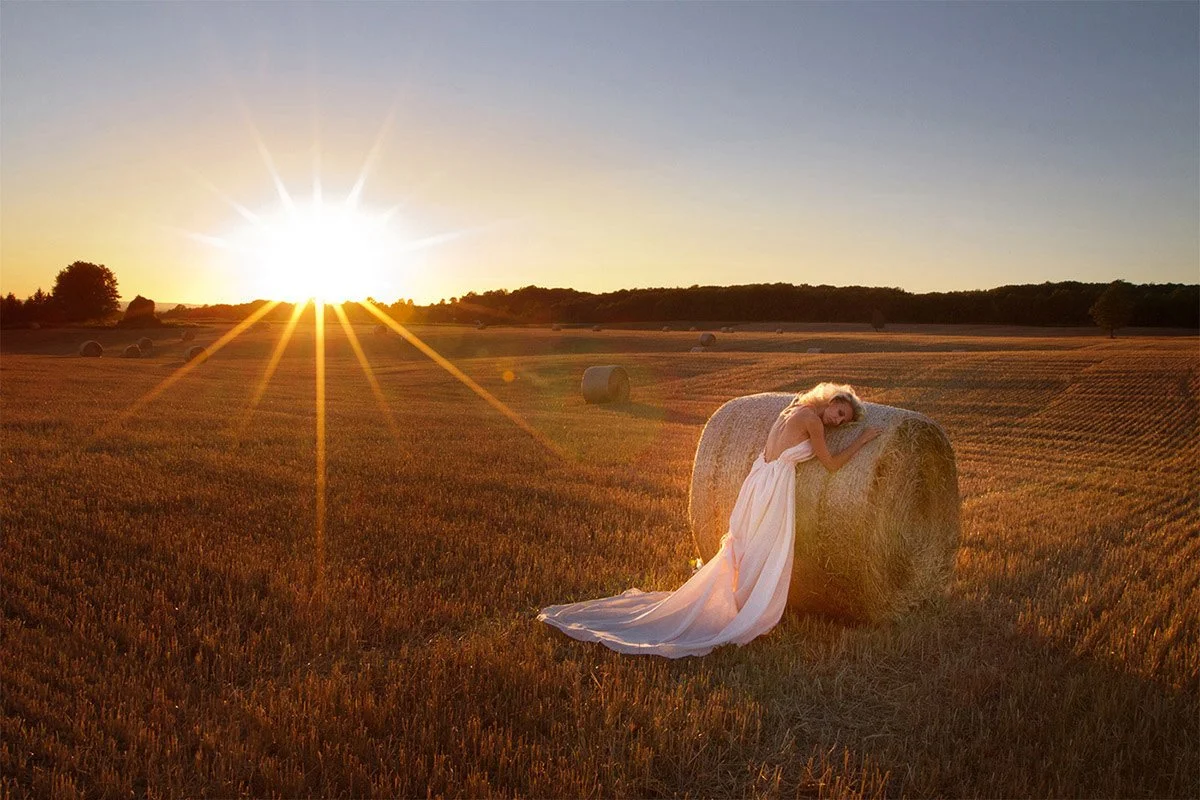 A woman in a white dress leaning on a large hay bale in a field during sunset.