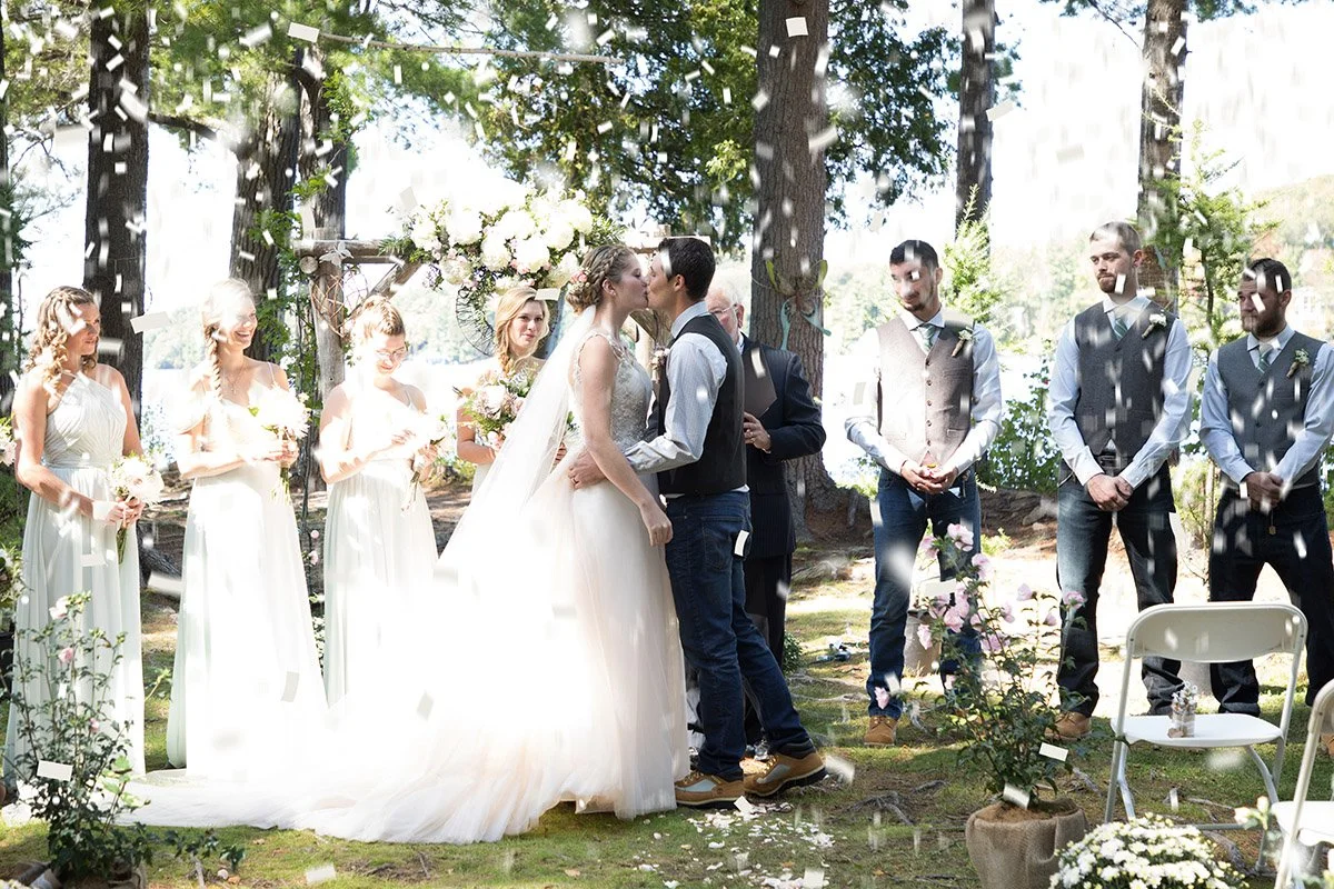 A couple sharing a kiss at their outdoor wedding ceremony, surrounded by bridesmaids and groomsmen, with confetti falling from above and trees in the background.