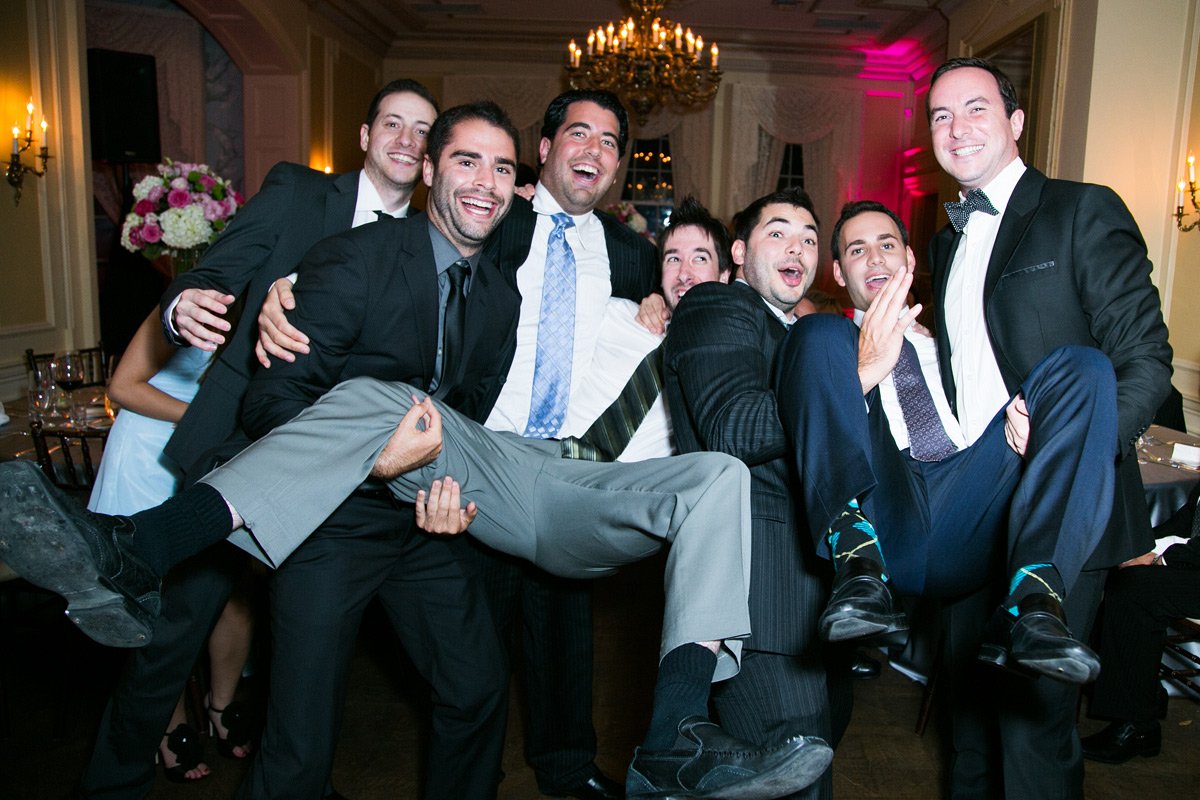 Group of men in suits at a formal event, lifting one man in the air, all smiling and laughing, in an elegant room with chandeliers and floral arrangements.