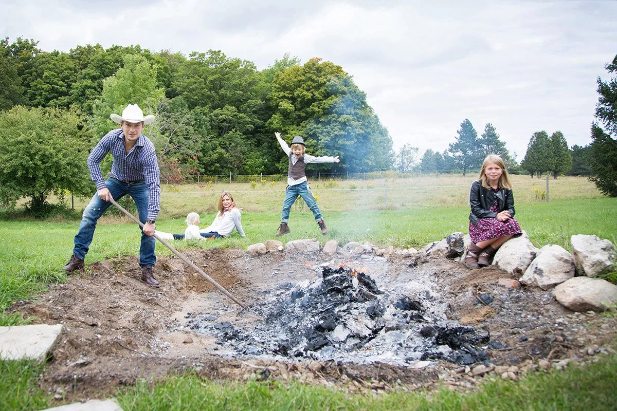 A man with a cowboy hat and plaid shirt is tending to a campfire with a tool while four children are nearby, one jumping mid-air, one sitting on rocks, and two sitting on the grass.