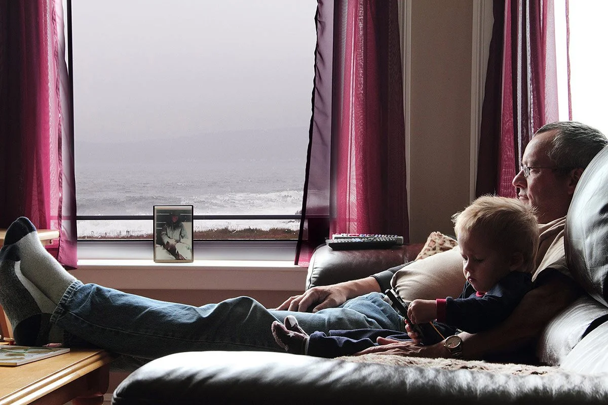 An elderly man and a young child sitting on a couch in a living room, watching the ocean through a large window with pink curtains.