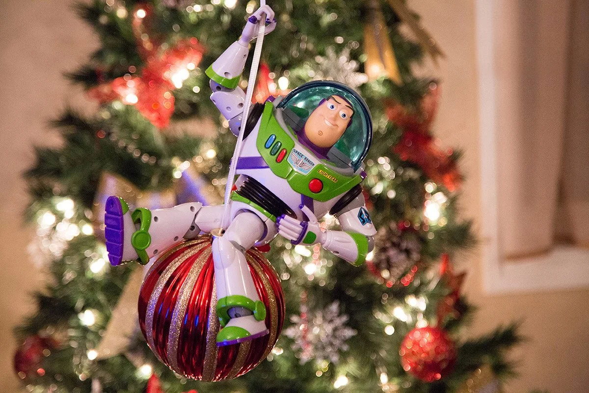 Buzz Lightyear toy hanging from a Christmas tree ornament, with Christmas lights and ornaments in the background.