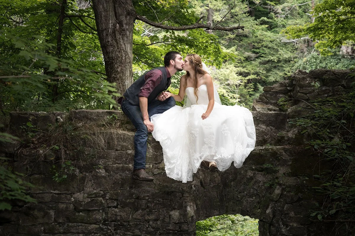 A couple in wedding attire sharing a kiss on a stone ledge in a forested area.