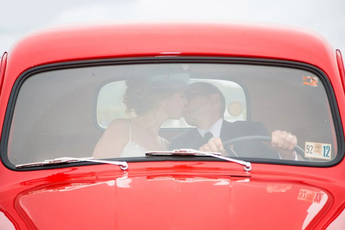 A bride and groom are sitting inside a red vintage car, kissing.