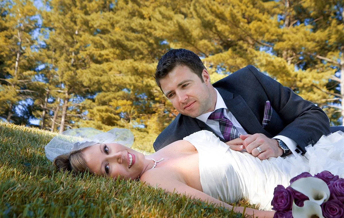 A bride lying on the grass in a white wedding dress with a veil, smiling, with a groom in a dark suit and purple plaid tie lying beside her, holding her hand, surrounded by green grass and trees with yellow leaves.