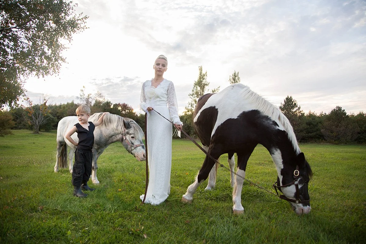 A woman and a young girl standing in a grassy field with two horses, one gray and one black and white, under a partly cloudy sky during sunset.