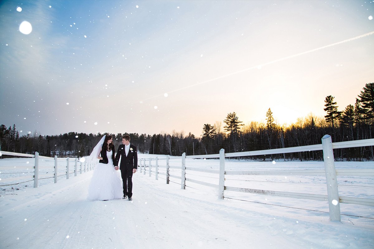 A bride and groom holding hands while walking on a snowy path in a winter landscape during sunset.