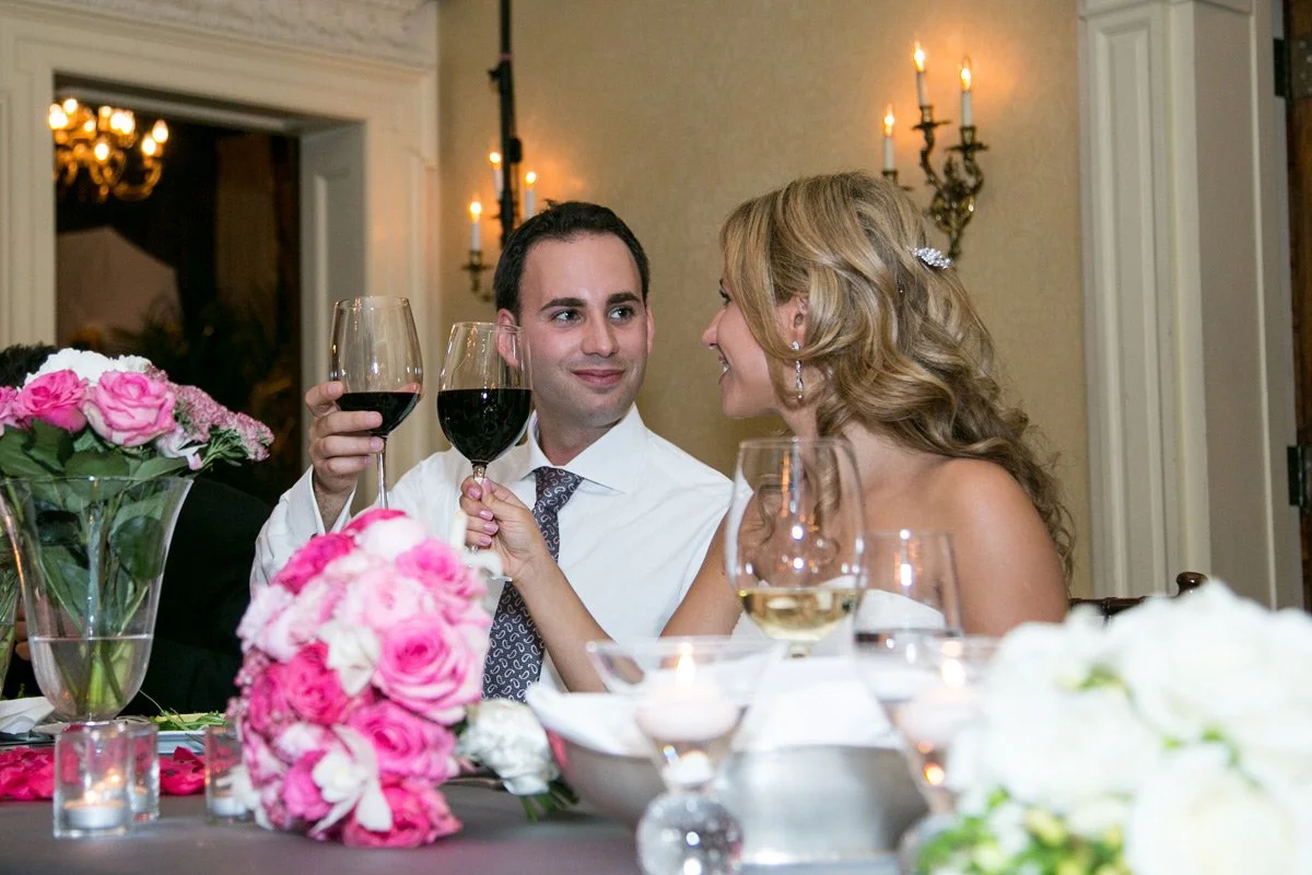 A man and woman at a formal dinner celebrating, holding glasses of red wine and smiling at each other, with floral centerpieces and candles on the table.