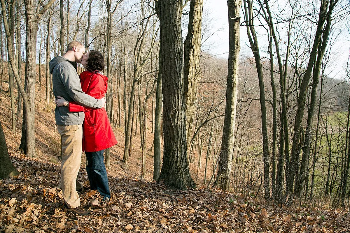 A couple kissing on a forest trail during fall, surrounded by leafless trees and fallen leaves.