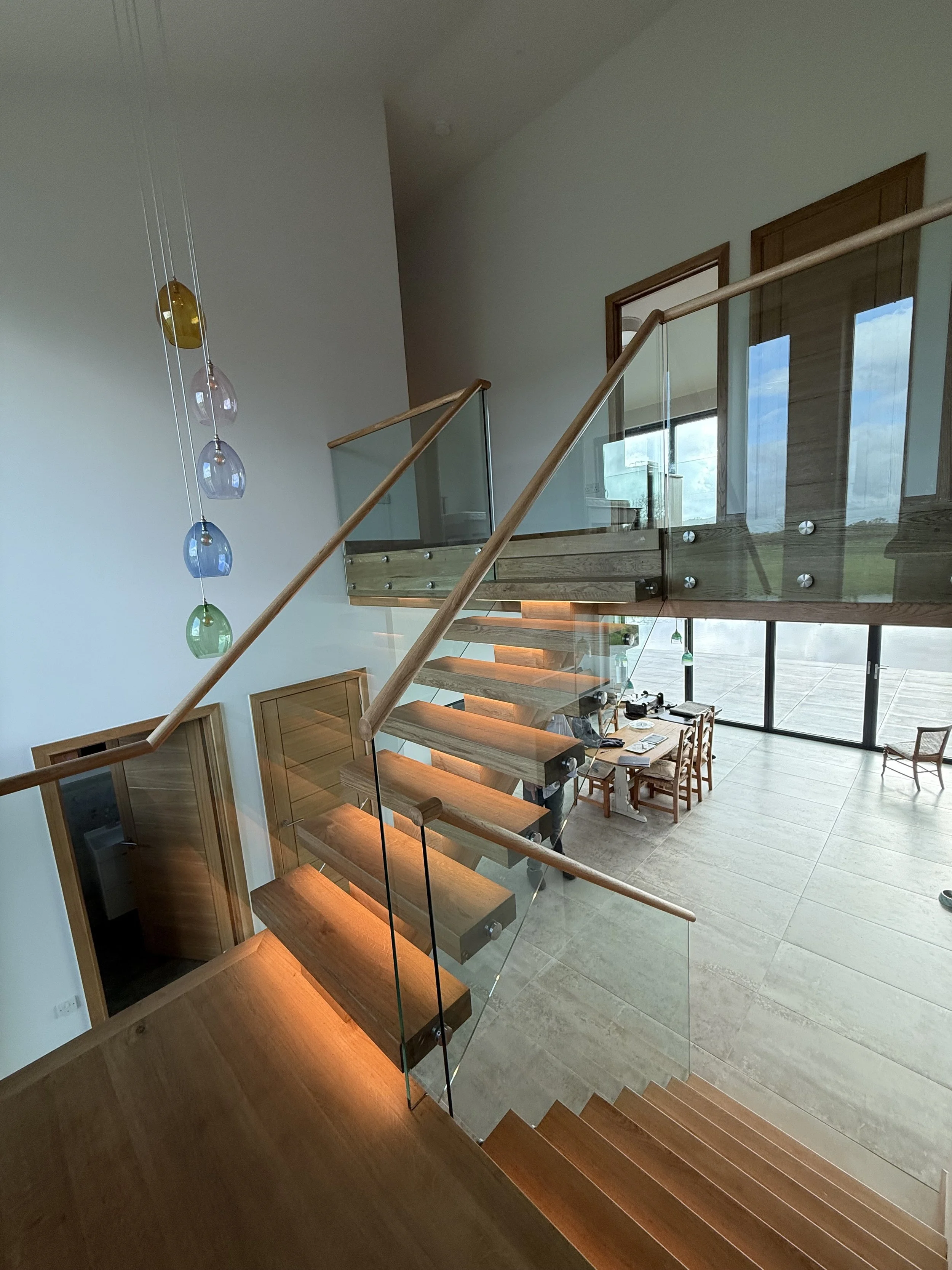 Interior view of a modern house with a staircase made of wood and glass railings, overlooking a high-ceiling living area with large windows letting in natural light, a dining table, and a decorative hanging light fixture.