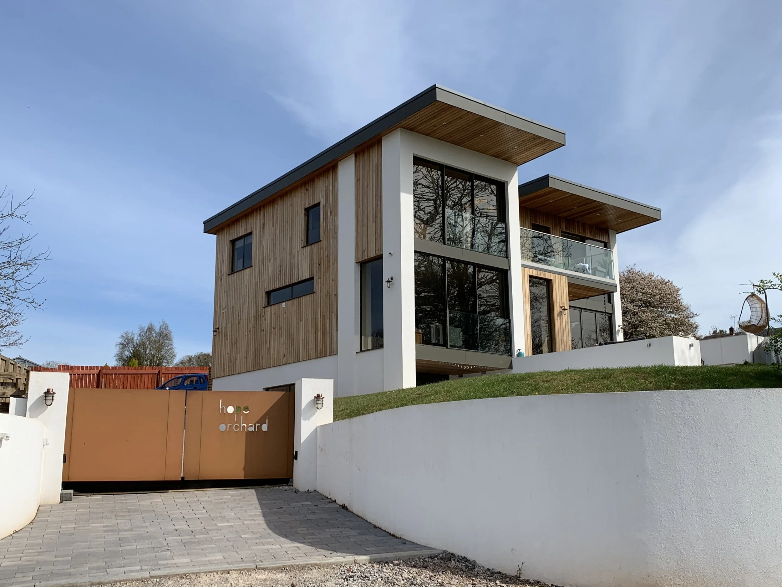 Modern multi-story house with cedar and white render exterior, large south facing glass windows and balcony, situated on a grassy hill behind a white wall and orange gate that says 'house orchard'.
