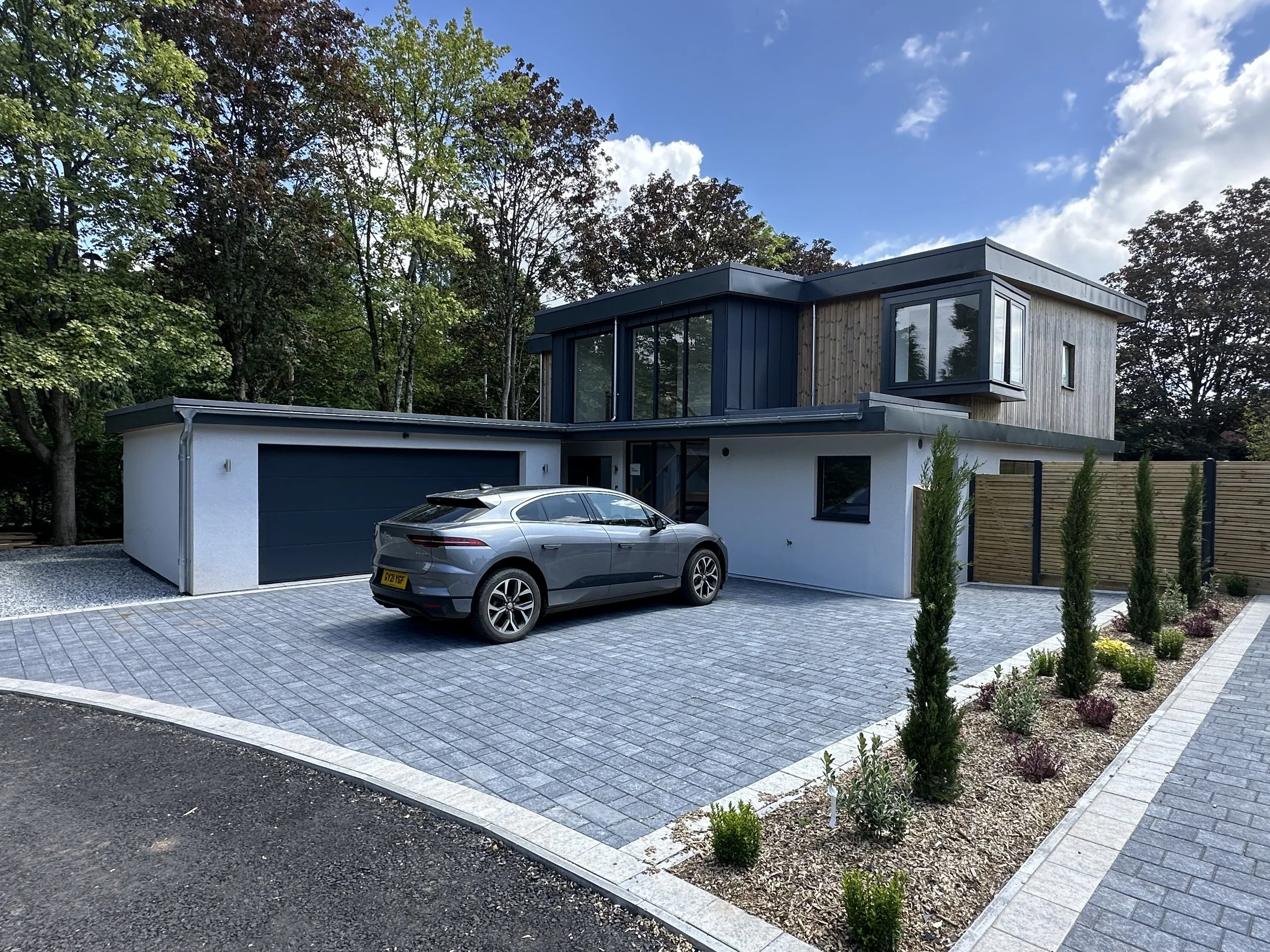 Modern house with white and wood exterior, a driveway with a gray car, and a landscaped garden with small trees and plants.