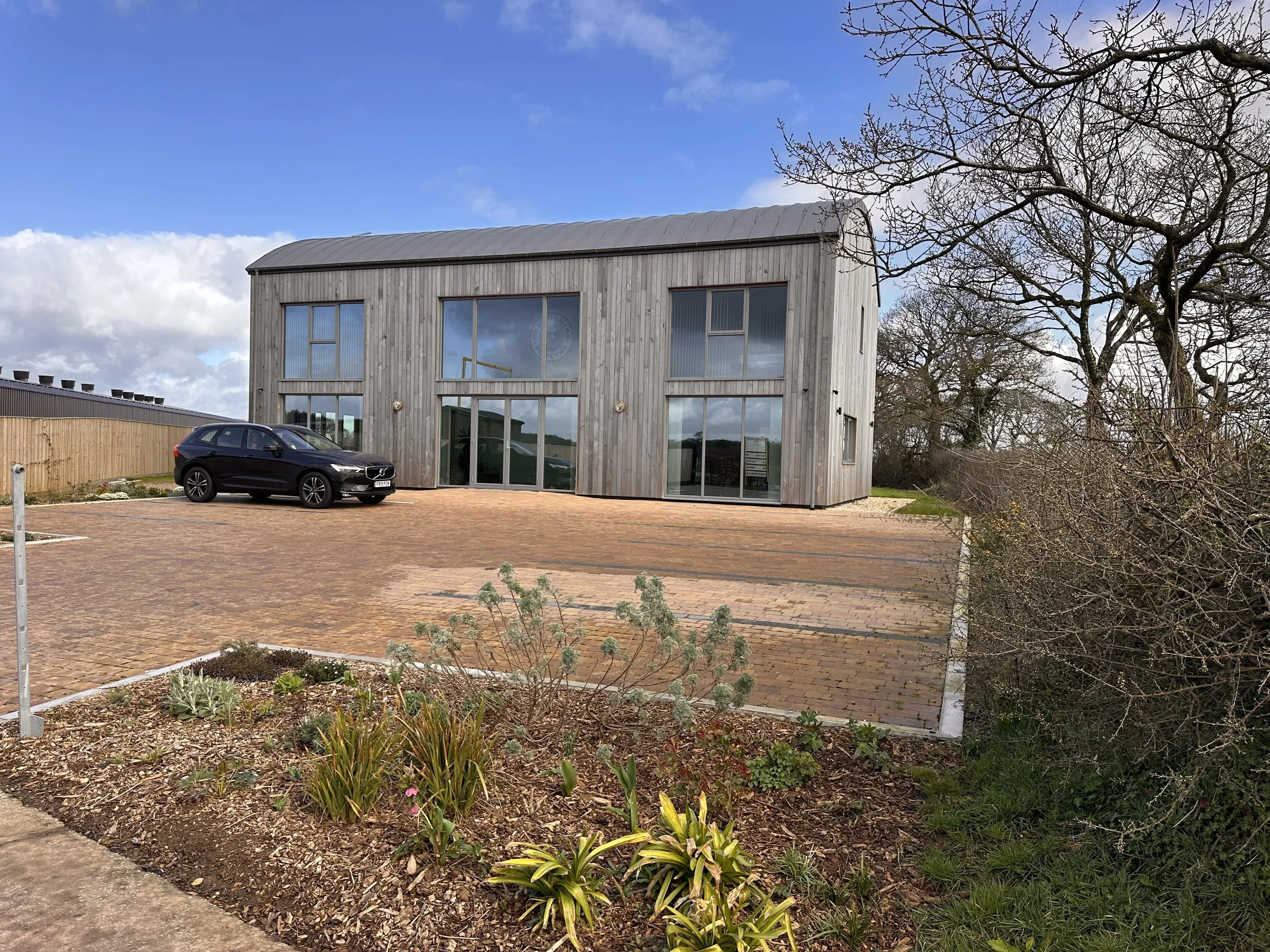 Modern wooden building with large glass windows, a black car parked outside on a brick-paved area, and a garden with plants in the foreground. It is a partly cloudy day with blue sky and some trees on the right side.