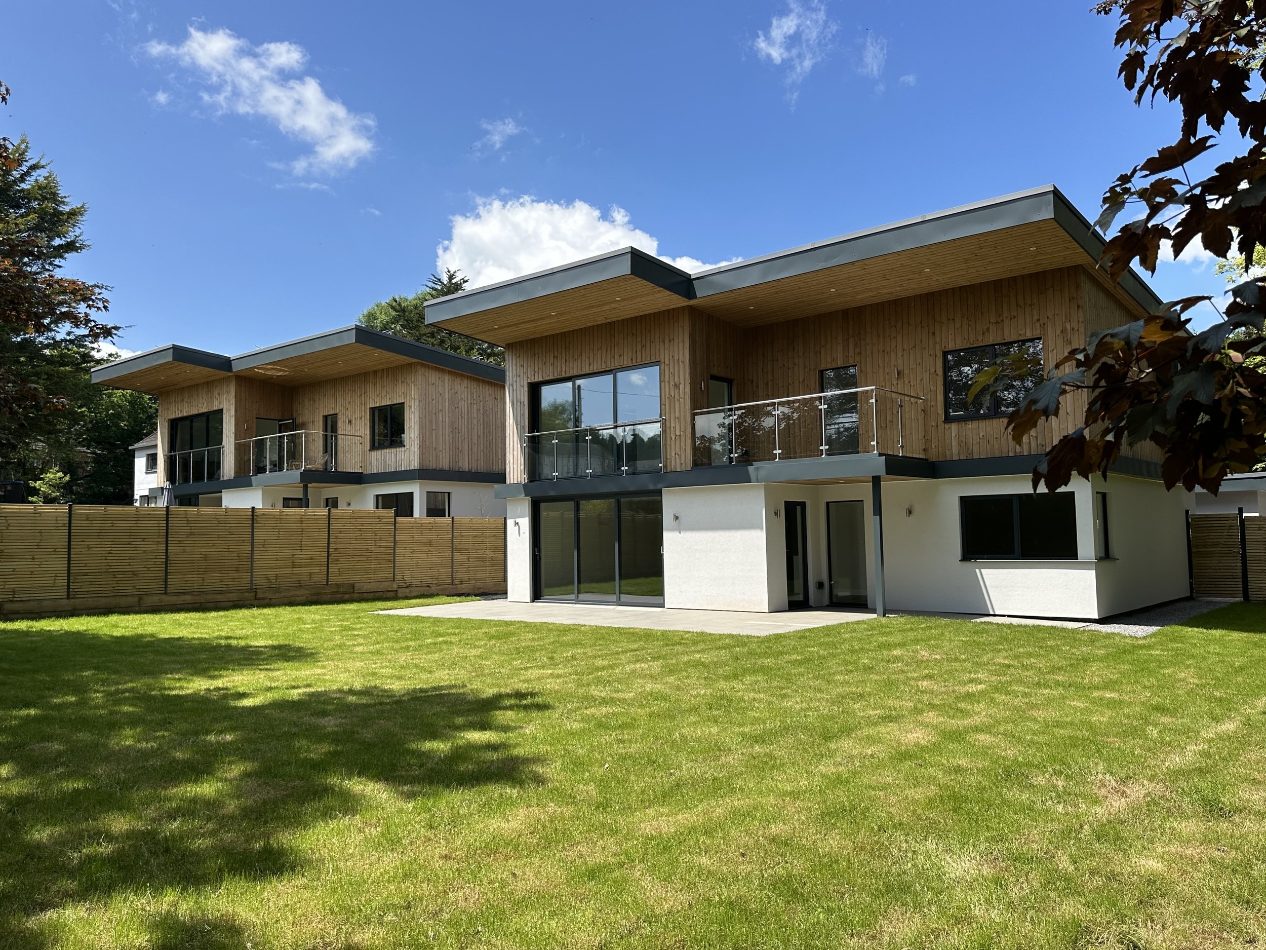 Modern two-story residential buildings with wood and white exterior, large windows, and balconies, surrounded by a grassy yard and a wooden fence, under a blue sky with clouds.