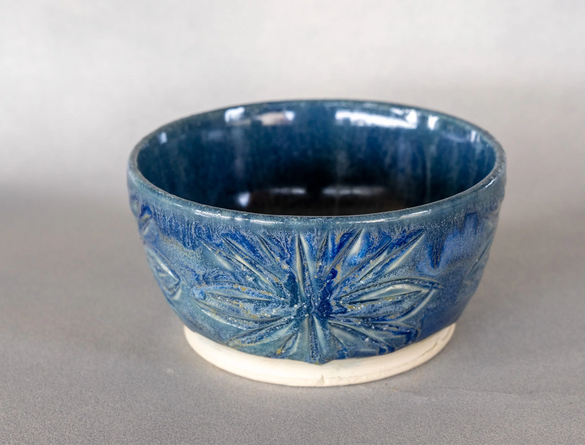A blue glass bowl with a textured floral pattern, placed on a white circular base on a gray surface.