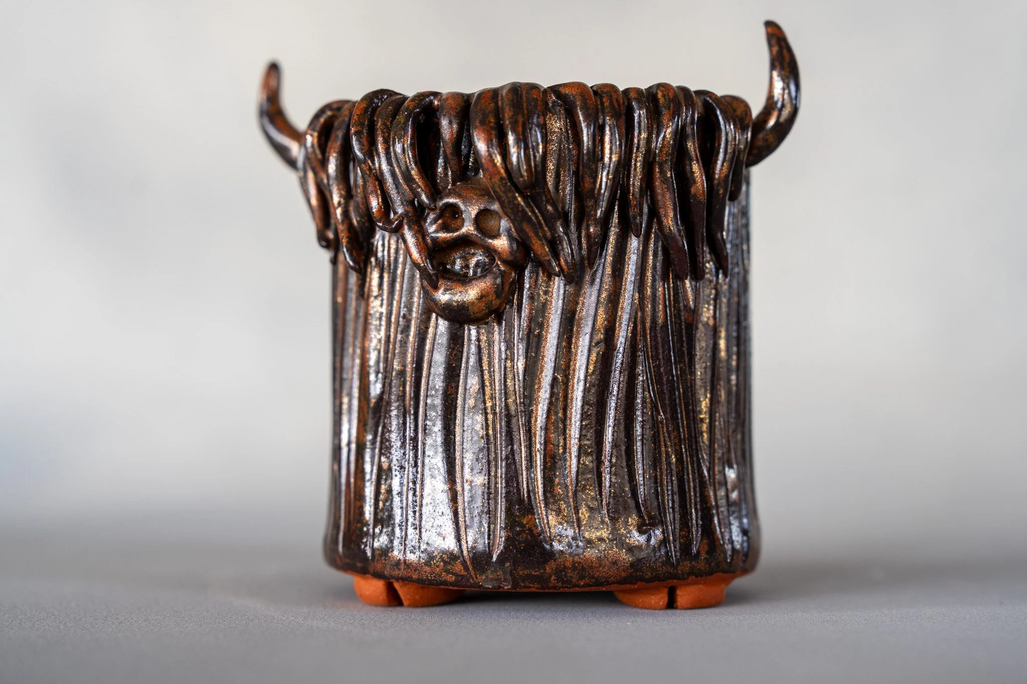 A ceramic sculpture of a stylized face with long hair and horns, with a glossy dark brown glaze and orange feet.