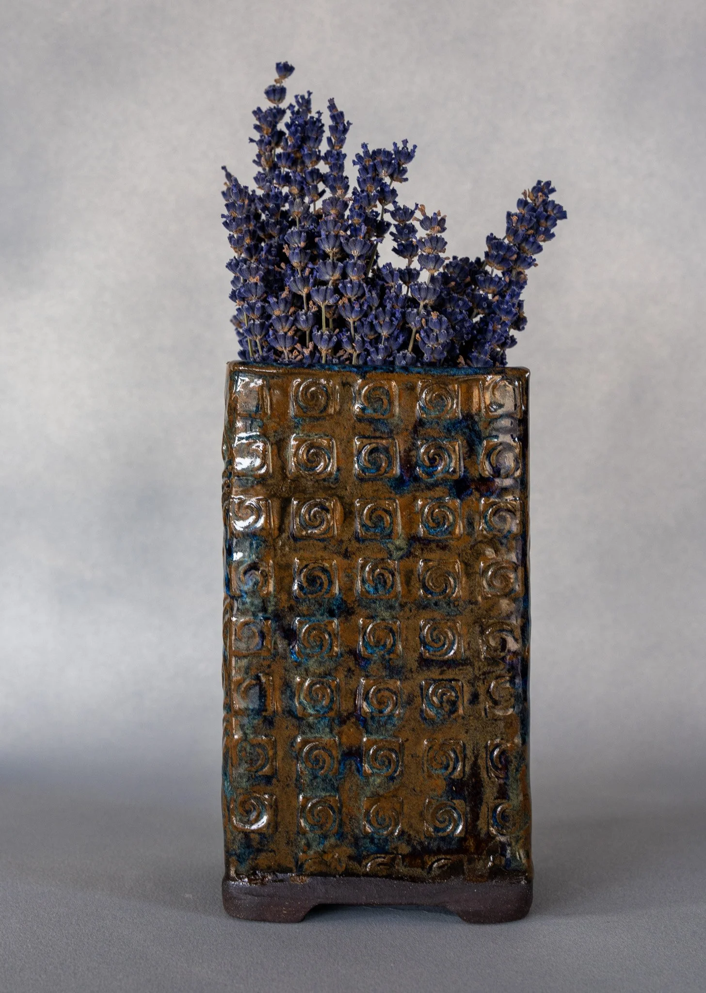 A ceramic vase with a textured, decorative pattern holding a bunch of dried lavender flowers.