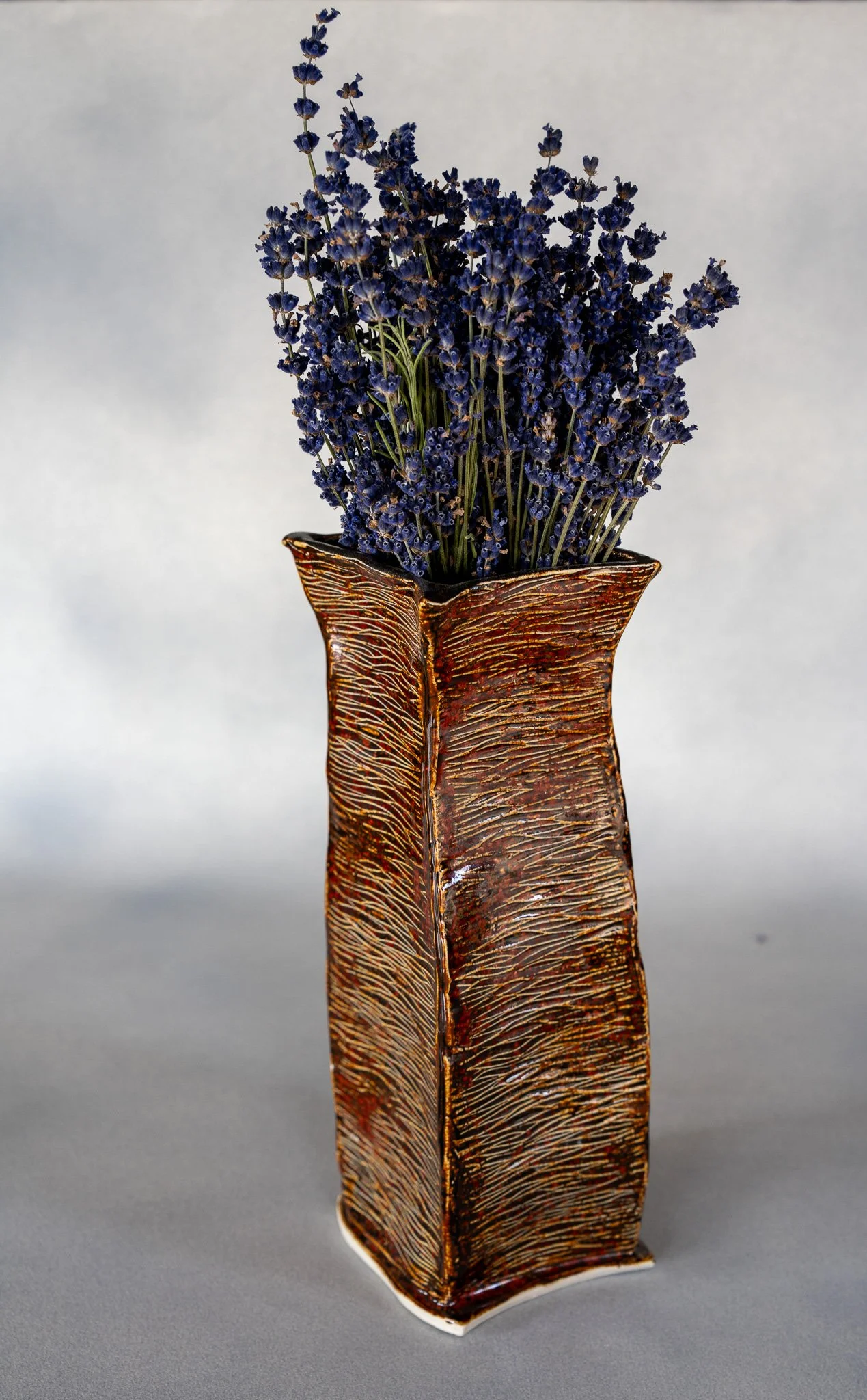 A decorative ceramic vase with a textured, brown and gold finish holds a bunch of dried lavender flowers.