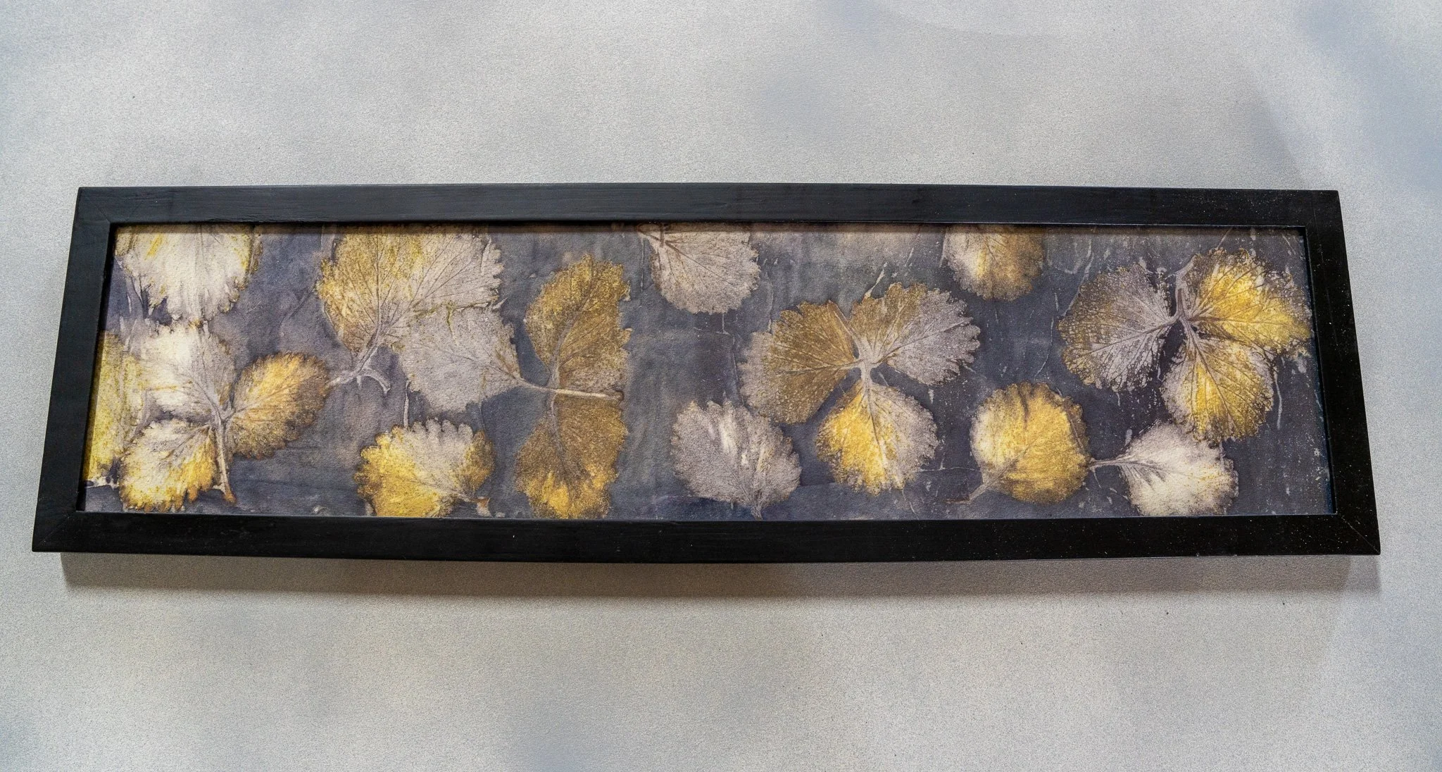 Framed artwork featuring pressed leaves in gold and silver tones on a dark background.